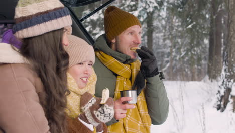 Side-View-Of-Parents-And-Daughter-Eating-Sandwiches-And-Drinking-Hot-Drink-Sitting-In-The-Trunk-Of-The-Car-In-A-Snowy-Forest