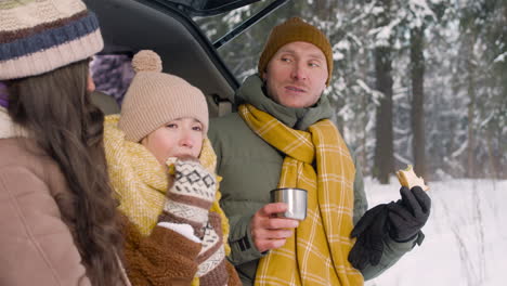Side-View-Of-Parents-And-Daughter-Eating-Sandwiches-And-Drinking-Hot-Drink-Sitting-In-The-Trunk-Of-The-Car-In-A-Snowy-Forest-1