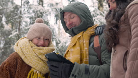 Bottom-View-Of-A-Father,-Mother-And-Daughter-Dressed-In-Winter-Clothes-Lookingt-At-Smartphone-In-A-Snowy-Forest