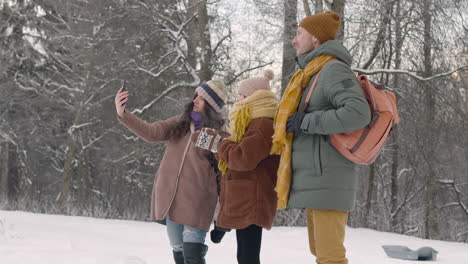 Father,-Mother-And-Daughter-Taking-Photos-With-Smartphone-In-A-Snowy-Forest