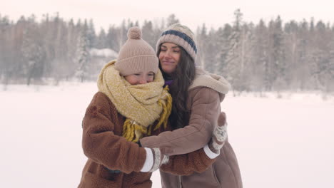 Front-View-Of-Mother-And-Daughter-In-Winter-Clothes-Hugging-And-Looking-At-Camera-In-A-Snowy-Forest