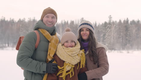 Front-View-Of-Father,-Mother-And-Daughter-In-Winter-Clothes-Hugging-And-Looking-At-Camera-In-A-Snowy-Forest