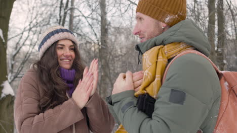 Husband-And-Wife-In-Winter-Clothes-Warm-Their-Hands-With-Their-Mouths-In-A-Snowy-Forest-1