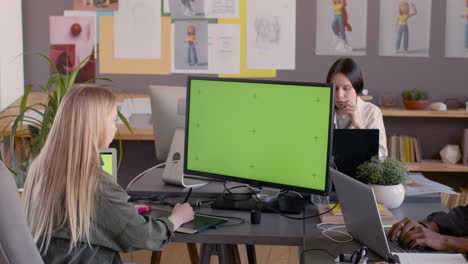 Female-Graphic-Designer-Using-Digital-Drawing-Tablet-And-Looking-At-Monitor-With-Green-Screen-In-An-Animation-Studio
