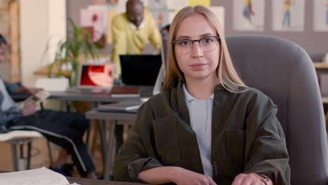 Portrait-Of-A-Smiling-Young-Woman-With-Eyeglasses-Looking-At-Camera-While-Sitting-At-Desk-In-An-Animation-Studio-And-Her-Multiethnic-Colleagues-Working-Behind-Her