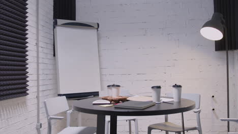 Empty-Small-Meeting-Room-With-Round-Black-Table,-White-Chairs,-Takeaway-Coffee,-Notebooks,-Floor-Lamp-And-Whiteboard