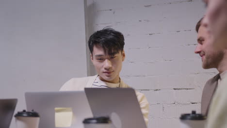 Young-Man-Looking-At-Laptop-Computer-Screen-And-Talking-With-Colleague-During-A-Team-Meeting