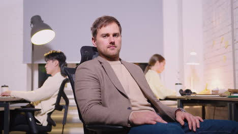 Portrait-Of-A-Confident-Businessman-Sitting-On-Office-Chair-And-Looking-At-Camera-While-Two-Employees-Working-Behind-Him