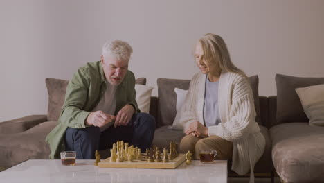 Senior-Mann-Und-Frau-Spielen-Schach-Und-Trinken-Tee,-Während-Sie-Zu-Hause-Auf-Dem-Sofa-Sitzen