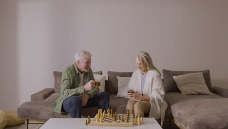 älterer-Mann-Und-Frau,-Die-Tee-Trinken-Und-Schach-Spielen,-Sitzen-Auf-Dem-Sofa-Zu-Hause