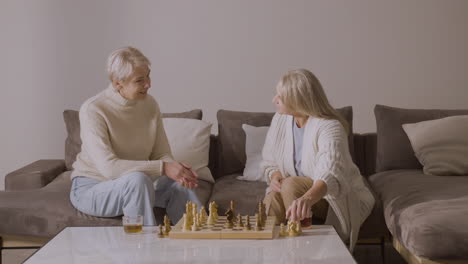 Two-Senior-Women-Playing-Chess-Sitting-On-Sofa-At-Home