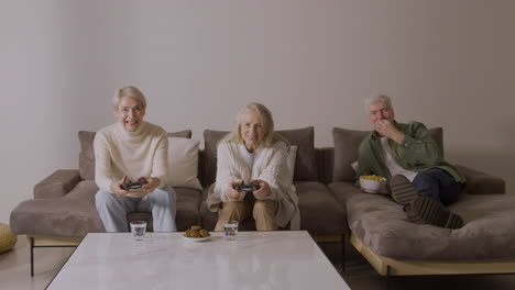 Zwei-Glückliche-ältere-Frauen,-Die-Videospiele-Spielen,-Sitzen-Zu-Hause-Auf-Dem-Sofa,-Während-Ein-Begeisterter-älterer-Mann-Auf-Dem-Sofa-Liegt,-Sie-Beobachtet-Und-Etwas-Isst