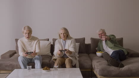 Zwei-Glückliche-ältere-Frauen,-Die-Zu-Hause-Auf-Dem-Sofa-Sitzen-Und-Videospiele-Spielen,-Während-Ein-Begeisterter-älterer-Mann-Auf-Dem-Sofa-Liegt-Und-Sie-Beobachtet-Und-Etwas-Isst-1