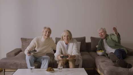 Zwei-Glückliche-ältere-Frauen,-Die-Zu-Hause-Auf-Dem-Sofa-Sitzen-Und-Videospiele-Spielen,-Während-Ein-Begeisterter-älterer-Mann-Auf-Dem-Sofa-Liegt-Und-Sie-Beobachtet-Und-Etwas-Isst-2
