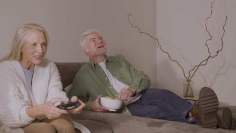 Glückliche-ältere-Frau,-Die-Videospiele-Spielt,-Sitzt-Zu-Hause-Auf-Dem-Sofa,-Während-Ein-Begeisterter-älterer-Mann-Sie-Unterstützt
