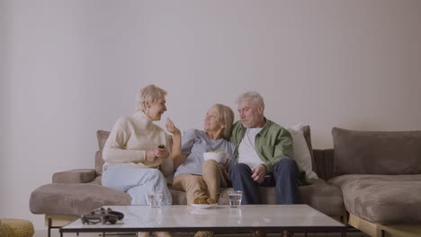 Gruppe-Von-Drei-Senioren,-Die-Fernsehen,-Reden-Und-Karamellpopcorn-Essen,-Während-Sie-Zu-Hause-Auf-Dem-Sofa-Sitzen