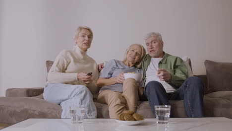 Gruppe-Von-Drei-Senioren,-Die-Fernsehen,-Reden-Und-Karamellpopcorn-Essen,-Während-Sie-Zu-Hause-Auf-Dem-Sofa-Sitzen-2