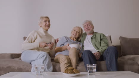 Gruppe-Von-Drei-Senioren,-Die-Fernsehen,-Reden-Und-Karamellpopcorn-Essen,-Während-Sie-Zu-Hause-Auf-Dem-Sofa-Sitzen-3