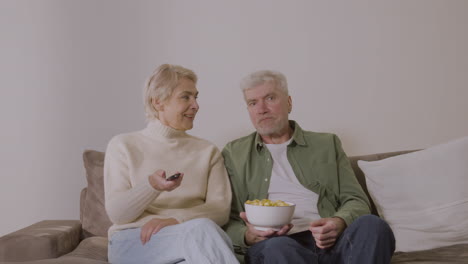 Senior-Couple-Talking-And-Watching-Tv-While-Sitting-On-Sofa-At-Home