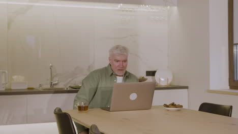 älterer-Mann-Sitzt-Auf-Einem-Stuhl-In-Der-Küche-Und-Benutzt-Einen-Laptop-Computer