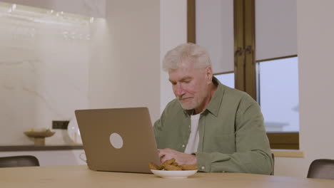 Glücklicher-älterer-Mann,-Der-Auf-Einem-Stuhl-In-Der-Küche-Sitzt-Und-Einen-Laptop-Benutzt