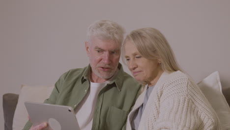 Senior-Couple-Sitting-On-Sofa-At-Home-Using-Digital-Tablet-1