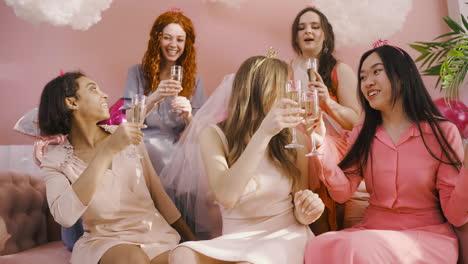 Vorderansicht-Einer-Gruppe-Von-Freunden-Und-Junggesellenmädchen,-Die-Champagner-Kristallgläser-Halten,-Während-Sie-Auf-Dem-Sofa-Sitzend-Tanzen