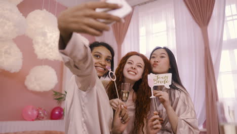 Three-Multiethnic-Friends-Holding-Cardboard-Decoration-And-Crystal-Glasses-Of-Champagne-Taking-A-Selfie-With-Smartphone-In-Bachelorette-Party