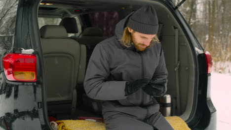 Handsome-Bearded-Man-Sitting-In-Car-Boot-And-Using-Mobile-Phone-On-A-Snowy-Winter-Day