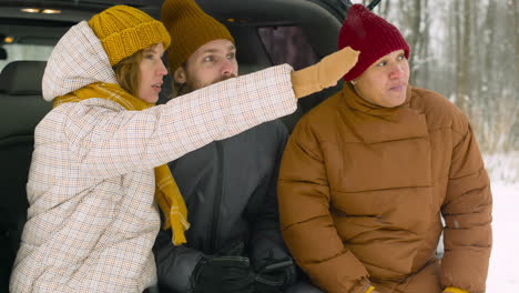 Three-Friends-Talking-And-Pointing-Something-While-Sitting-In-Car-Boot-On-A-Snowy-Winter-Day
