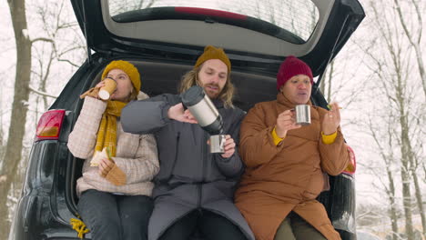 Three-Friends-Drinking-And-Eating-Sandwich-While-Sitting-In-Car-Boot-During-A-Winter-Road-Trip
