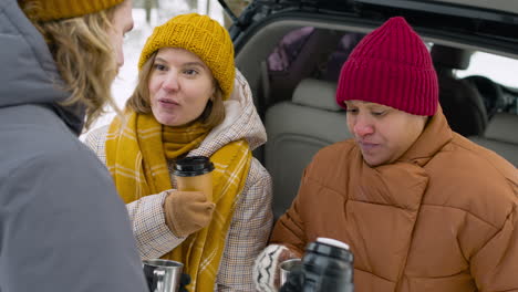 Cheerful-Friends-Drinking-And-Talking-Together-Near-Car-In-A-Snowy-Forest-During-A-Winter-Road-Trip