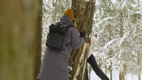 Couple-In-Winter-Clothes-Playing-And-Hiding-Behind-A-Tree-Trunk-In-Winter-Forest