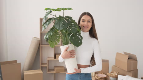 Happy-Woman-Walking-With-Potted-Plant-And-Smiling-At-Camera-At-New-Home