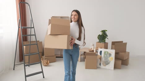 Happy-Woman-Holding-Boxes-And-Walking-In-Empty-Apartment-While-Smiling-At-Camera