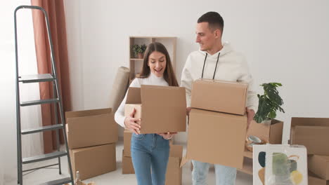 Happy-Couple-Moving-To-New-Place-Holding-Cardboard-Boxes-And-Walking-Together-In-Empty-Flat