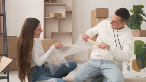 Zoom-In-Shot-Of-Young-Couple-In-A-New-House-Sitting-On-The-Floor-Unrolling-Bubble-Wrap