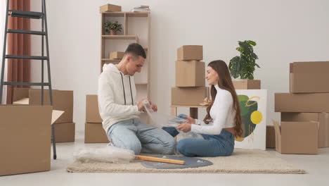 Zoom-In-Shot-Of-Young-Couple-In-A-New-House-Sitting-On-The-Floor-Unrolling-Bubble-Wrap-1