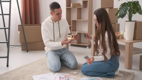 Young-Couple-In-A-New-House-Sitting-On-The-Carpet-And-Choosing-Colours-For-Decoration-Wth-A-House-Model