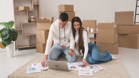 Young-Couple-In-A-New-House-Sitting-On-The-Carpet-With-Laptop-Choosing-Colours-For-Decoration
