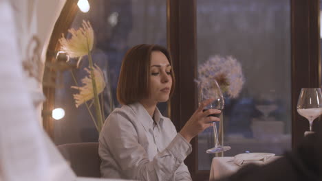 Serious-Woman-Sitting-Alone-At-Restaurant-Table-And-Drinking-Wine-While-Female-Musician-Playing-Piano