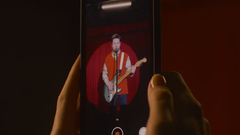 Close-Up-View-Of-Smartphone-Held-By-Hands-Recording-A-Male-Musician-Playing-Guitar-And-Singing-During-Live-Music-Perfomance