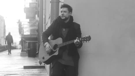 Black-And-White-View-Of-A-Guitar-Case-On-The-Floor-And-A-Man-In-Coat-Singing-And-Playing-Guitar-Leaning-On-The-Wall-In-The-Street
