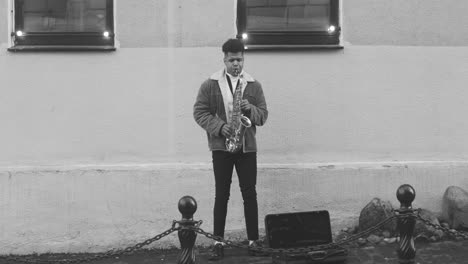 Black-And-White-And-Front-View-Of-A-Sax-Case-On-The-Floor-And-A-Man-In-Jacket-Playing-Sax-In-The-Street