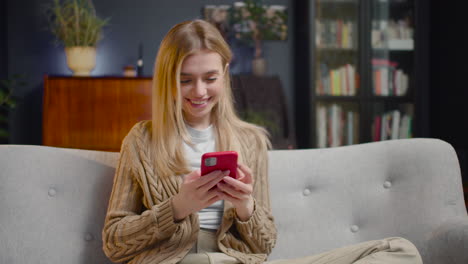 Happy-Young-Woman-Chatting-On-Smartphone-And-Laughing-While-Sitting-On-A-Couch-At-Home