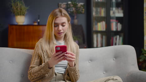 Smiling-Young-Woman-Using-Smartphone-While-Sitting-On-A-Couch-At-Home