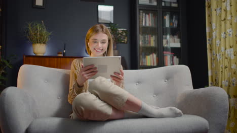 Smiling-Young-Woman-With-Earphones-Using-Tablet-While-Sitting-On-Couch-At-Home