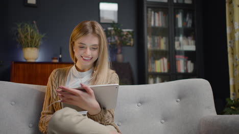 Smiling-Young-Woman-With-Earphones-Using-Tablet-While-Sitting-On-Couch-At-Home-1