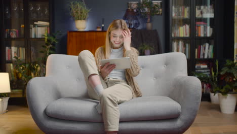 Happy-Young-Woman-With-Earphones-And-Tablet-Entering-Living-Room-And-Sitting-On-Couch