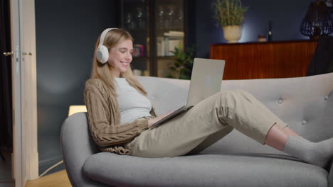 Happy-Young-Woman-With-Wireless-Headphones-Chatting-On-Laptop-Computer-While-Lying-On-Couch-At-Home-1